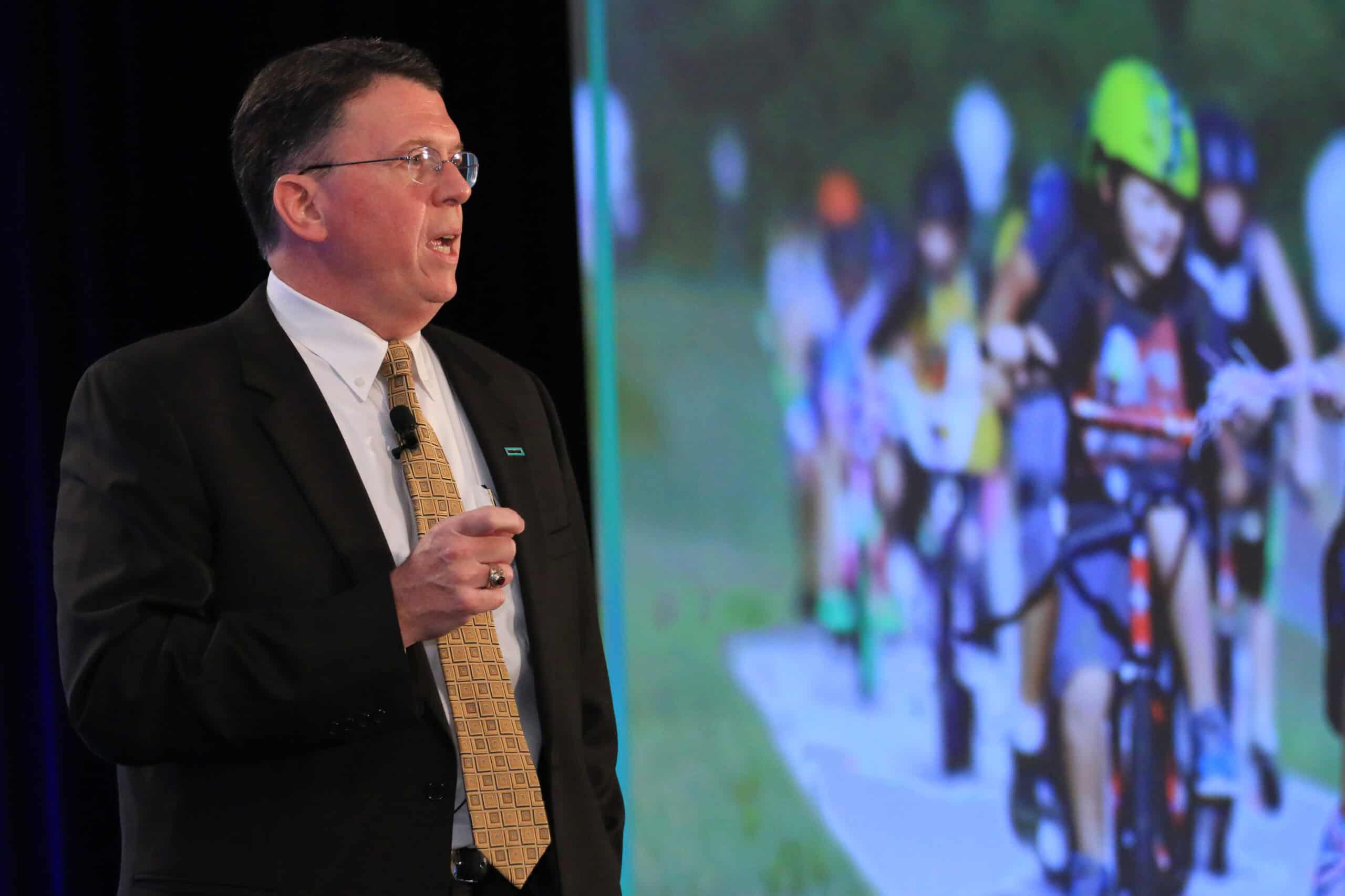 Conference attendee speaking with a slide in the background of children on bikes